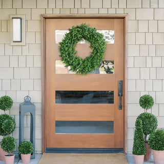Wooden front door with a green wreath, flanked by potted plants on a tiled wall background.