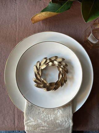 An elegant setting showcasing a single 7.5" Iron Wreath Ornament in brass by CG Hunter, placed atop a white plate on a textured table runner, exuding a luxurious holiday charm.