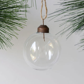 Timeless 2.25" Clear Glass Ornament by CG Hunter, elegantly hanging against a backdrop of lush pine branches, reflecting light and enhancing holiday decor.